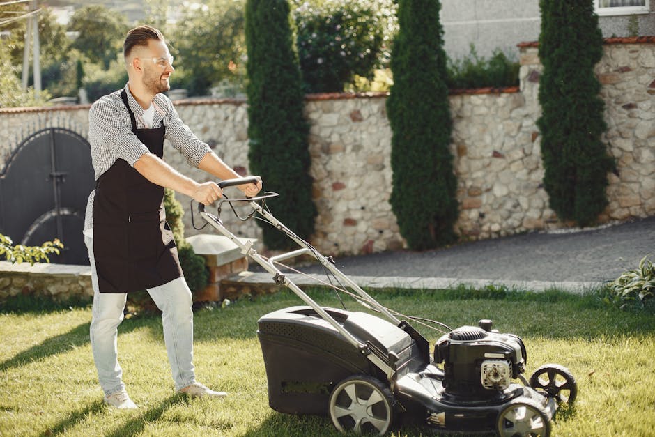 A man wearing a black apron over a checkered shirt and light-colored pants is pushing a lawn mower across a well-maintained garden. The lawn mower is black with a visible engine and large rear wheels. The garden features lush green grass, bordered by a stone wall with mortar, topped with red tiles, and complemented by tall, narrow conifer trees and leafy shrubs. In the background, there's a gravel pathway beside the wall, and some sunlight filters through the trees, casting gentle shadows. The scene depicts active garden maintenance, with the man engaged in lawn care work. The setting appears tidy and cared-for, aligned with professional landscaping work typically conducted by Gardeners Balham, and emphasizes the outdoor maintenance theme suitable for homeowners.